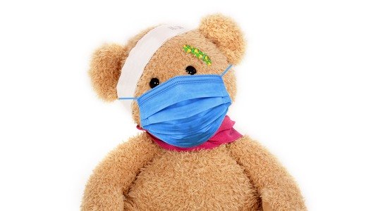 Ein brauner Teddy hat ein grünes Pflaster auf der Stirn und einen weißen Verband um seinen Kopf. Er trägt einen blauen Mundnasenschutz.